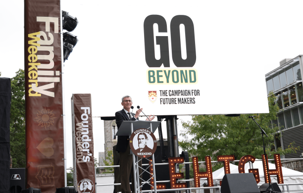 President Joseph J. Helble stands at a podium clapping with the GO Beyond signs and Family and Founder's Weekend banners.