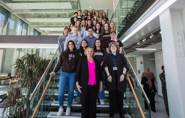 Dean Dolan poses on stairs in the Health, Science, and Technology building with a large group of college of health students standing on the stairs behind her