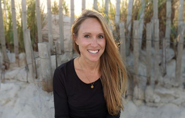 Deidre Martin wears a black long sleeved shirt and seated in front of the dunes on a beach smiling at the camera.