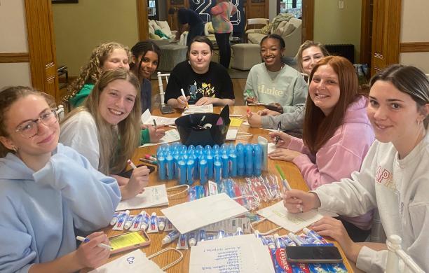 Sorority sisters sit with high school Girl Scouts at a table covered with toothpaste, toothbrushes, and deodorant to create care packages.