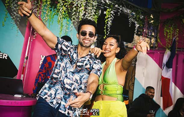 Aakash Phulwani in a loud shirt and dark sunglasses points skyward as he dances with a woman in neon
