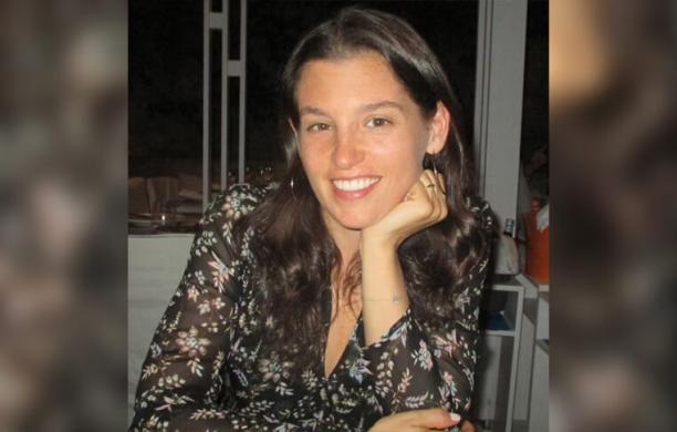 Woman with brown hair smiles and poses while sitting with her elbows on the table, resting her face on one hand