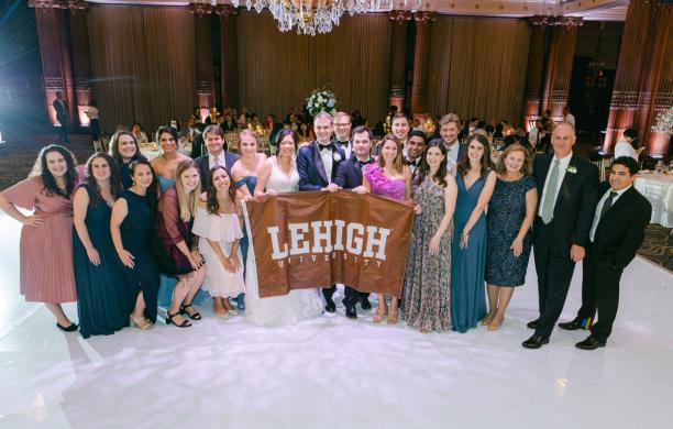 A large group of people surrounding Becky Lok and Mike Meehan at their wedding reception, holding a Lehigh flag.