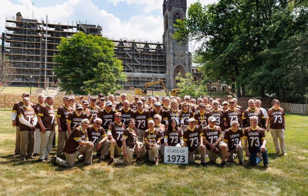 Alumni wear brown "73" Lehigh jerseys and pose in front of the University Center construction site holding a white sign that says "Class of 1973."