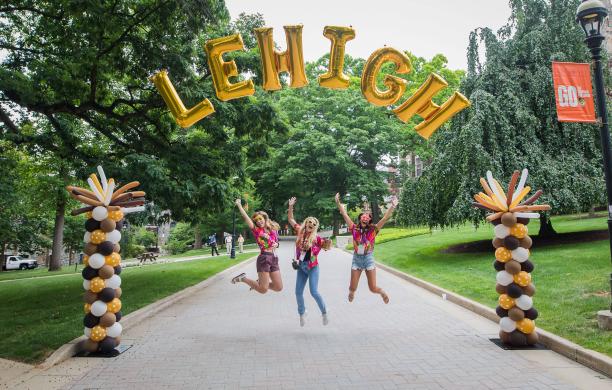 Three alumnae dressed in bright clothes jump with their arms in the air under an arch of balloons spelling out "Lehigh"