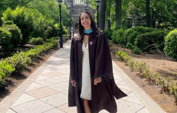 A smiling new graduate stands on a campus walkway wearing her graduation gown and a white spring dress.