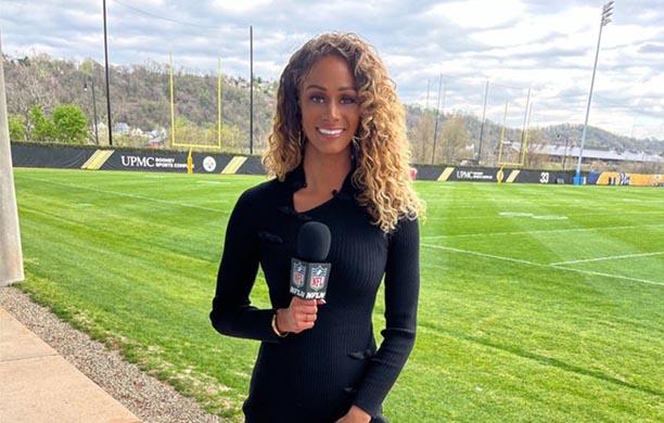 Kayla Burton holding a microphone and standing before a yellow goalpost on a lush, green football field.