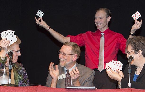 Philip La Porta in a red shirt holds up cards to the delight of the three people in front of him watching a magic trick