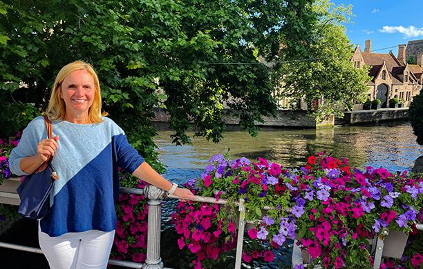 Diane Frisch smiles next to a display of purple flowers and a river in Bruges