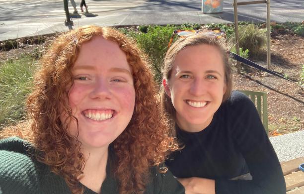 Two women pose for a selfie while sitting outside on a park bench and smiling.