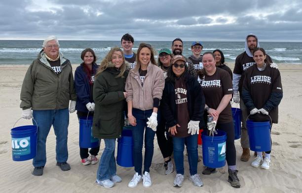 13 alumni carry large buckets and wear their brown and white with cold weather gear on the beach in front of the ocean shoreline.