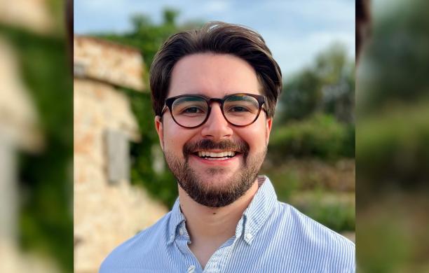 Peter Luba smiles at the camera outside with blue skies and stone building in the background