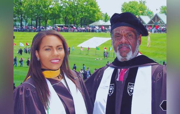 Both dressed in commencement regalia, a man stands beside a female graduate with a field behind them.