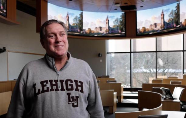 Bill Glaser wearing gray sweatshirt with Lehigh in brown stands smiling with images of the reimagined University Center behind him.