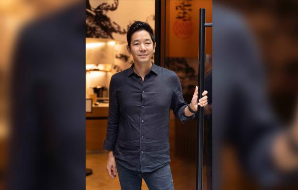 Man stands in the doorway of his restaurant, wearing a dark navy button-down and holding the door ajar with a slight smile.