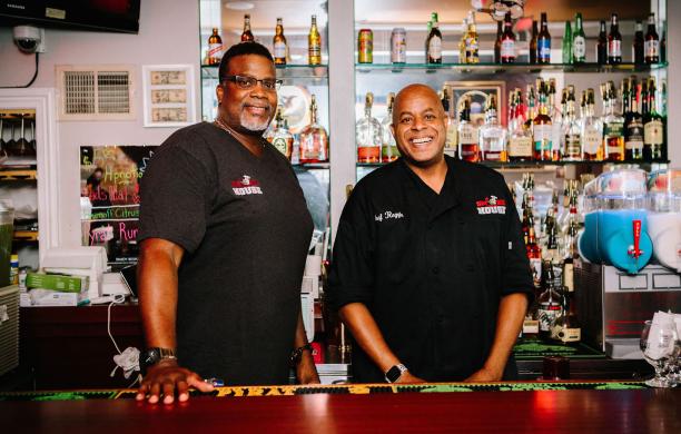 Two men, one with "Chef Reggie" on his shirt, stand behind a bar wearing black shirts with the Smokehouse BBQ logo.