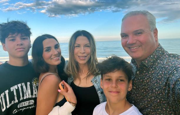 Family of five stands huddled for a selfie on a beach with the water, blue skies, and clouds behind them.