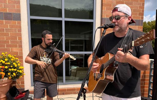 Both outdoors, one musician wears a brown Lehigh baseball shirt and plays a violin; another plays a guitar at a microphone.
