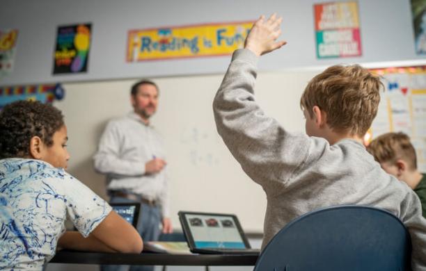 In a classroom, two students face a teacher standing at a whiteboard, one student raising his hand.