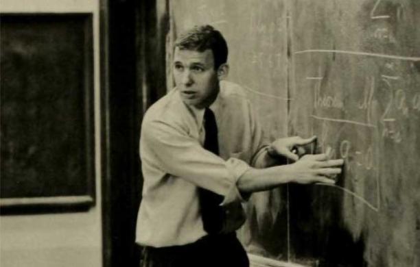 A professor gesturing at writing on a chalk board.