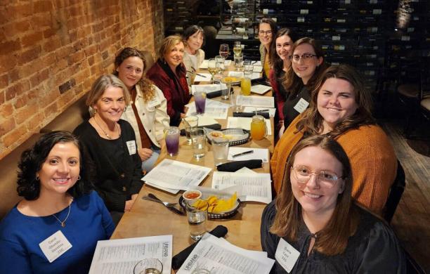A group of women sit at a long dining table with menus in front of them, posing for the camera.