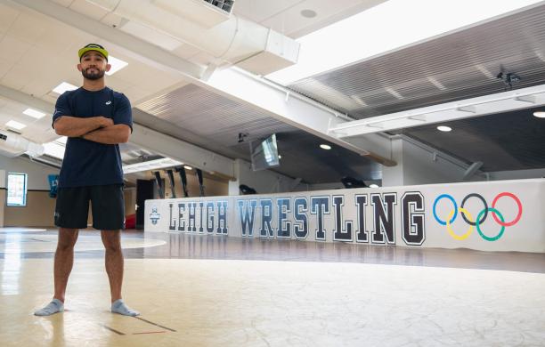 Darian Cruz stands with arms crossed on the mat in Grace Hall with the words "Lehigh Wrestling" and the Olympic rings behind him.