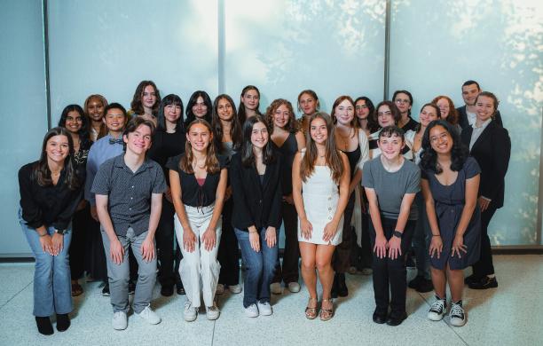 Standing in 3 rows with the front row squatting with hands on their knees, Students smile while posing against a wall of windows. 