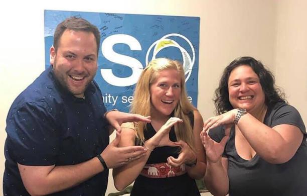 Three people pose beside each other posing in front of a CSO banner, each one holding up their hands to spell C - S - O.