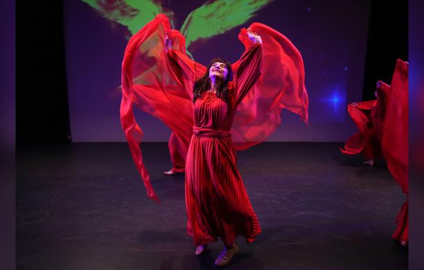 Female performer in a flowing red costume dances on a dark stage with sleeves floating in the air above her head.