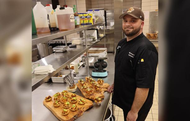 Man wearing a dark chef's coat and brown Lehigh hat stands in a commercial kitchen beside a stainless steel counter covered with appetizers.
