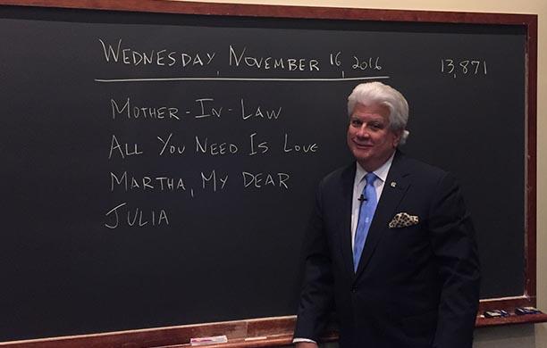 CJ Skender '76 stands in a suit in front of a blackboard with four music trivia clues