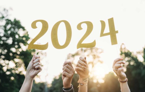 four hands of varying pigmentation holding golden and glittering numbers on sticks. The numbers make up "2024."