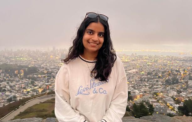 Forum Patel sits on a stone wall above the city of San Francisco with small building in the distance behind her.