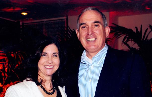 Sherry Hay stands beside Lew Hay in a dimly lit room, both dressed in business casual attire.