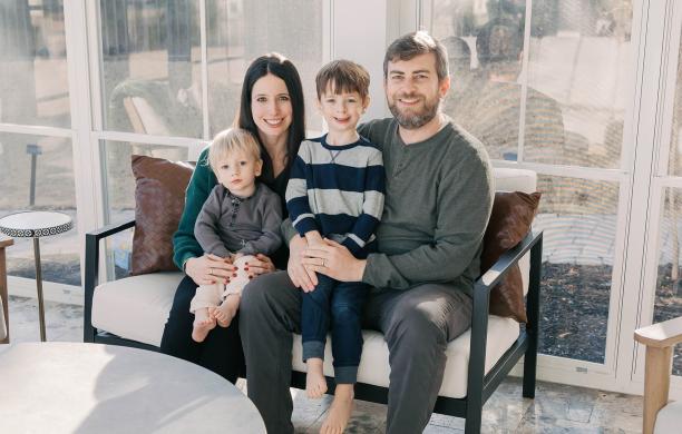 Family sits for a portait in a well lit white room against a wall of windows, with father and mother each holding a small boy in their lap.