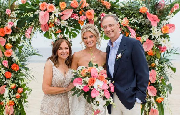 Fran & Tom Bartlett in formal wear on the beach beside their daughter in a wedding gown and carrying pink and peach flowers under a matching floral garland.