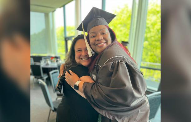 Carolina Hernandez side hugs a female student wearing a brown graduation cap and gown.