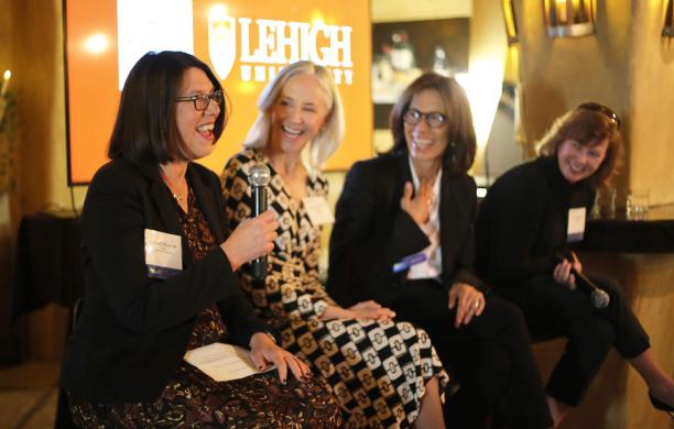 Four female panelists sit on bar height stools, the first speaking with a microphone while the others look on, smiling and laughing.