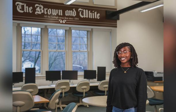 Emmanuella Agyemang stands in room, smiling in a black long sleeved shirt with a banner reading “The Brown and White” hangs behind her.