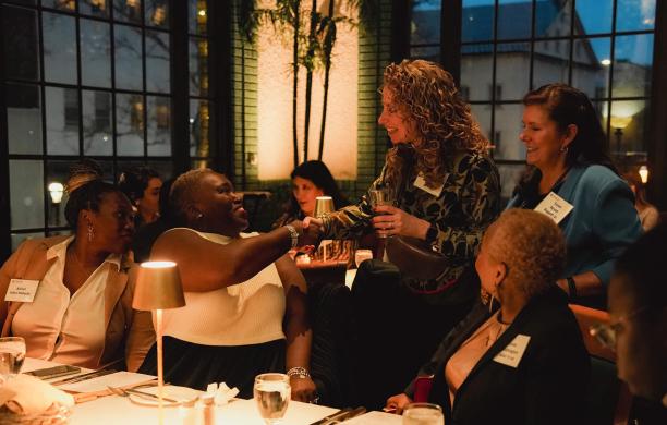 A table of women sitting with drinks in front of them turn to greet two other women approaching them and the center seat shakes hands with one.