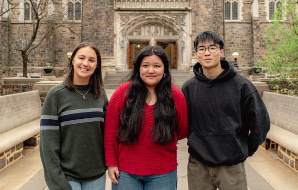 Standing in front of Alumni Memorial, 3 students stand side by side, posing with smiles for a photograph.