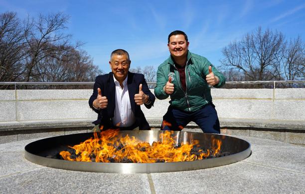 Gary and Garrett Pan stand behind the blazing fire pit, squatting behind it and each giving the camera a double thumbs up.