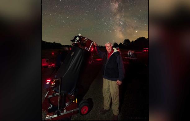 Man stands beside a large telescope in an open space on a dark night with starry skies clearly showing the Milky Way Galaxy above him.