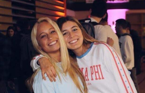 One college woman leans into a hug with another similarly aged as they pose in an auditorium with one arm around the other.
