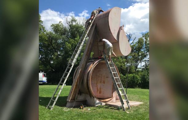 Two ladders stand against either side of a rusty sculpture while men on each work to refurbish it.