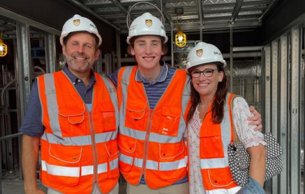 Doug, George, and Elissa Tiesi stand side-by-side, wearing orange construction vests and hardhats inside a construction site.