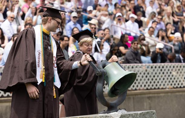 Both wearing brown graduation caps and gowns, a tall male student and an alumna from the class of 1975 both hold a large bell to ring it with a crowd in the stands behind them.