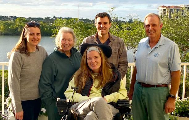 Family poses on a balcony over a lake, with Brooke Eby seated center and sister, mother, brother, and father standing behind her.