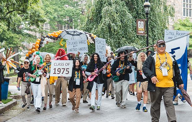 A group of alumni parade with long wigs and signs that read "Class of 1975" and "Still Rockin' and Rollin”
