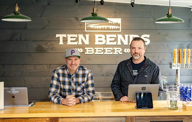Mike Scarlata and Jason Powell stand at a light wood counter with the Ten Bends Beer logo on the wall behind them